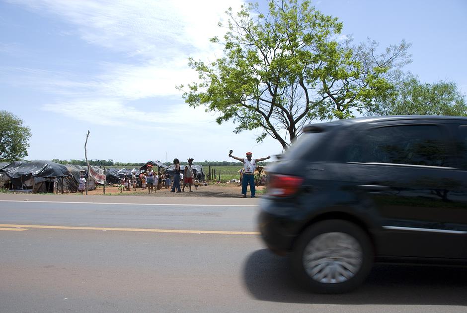 Un hombre guaraní de pie junto a una polvorienta carretera, con los brazos extendidos y una maraca mbaraka en su mano izquierda.

La deforestación de Mato Grosso do Sul ha obligado a muchos guaraníes, los propietarios originarios de la selva, a hacinarse en pequeñas parcelas de terreno.

Ya no existen, en su mayoría, los huertos en los que plantaban mandioca (o yuca) y maíz. También ha desaparecido la posibilidad de cazar libremente. En vez de eso, ahora están rodeados de ranchos de ganado, campos de soja y plantaciones de caña de azúcar.

El impacto de la pérdida de sus tierras sobre los guaraníes es muy profundo. _“Te vuelves espiritualmente vacío”_, confiesa un hombre guaraní.
