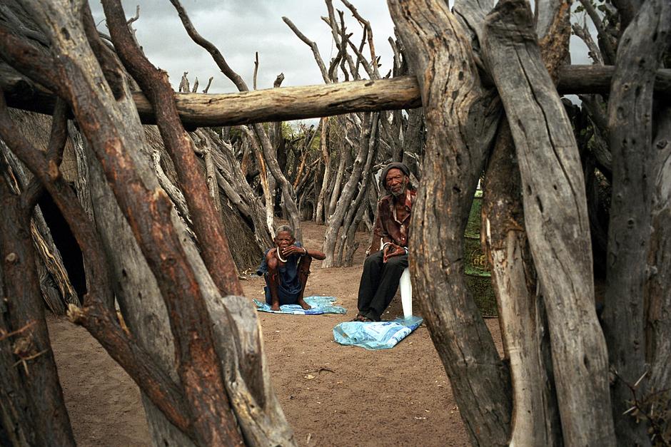 Un couple âgé de Bushmen dans leur terrain clos à New Xade, un camp de relocalisation dans lequel de nombreux Bushmen ont été déportés au cours des dix dernières années.

Au cours des trois grandes expulsions de 1997, 2002 et 2005, la plupart des Bushmen ont été forcés de quitter la réserve, en violation avec la constitution botswanaise.

Le gouvernement a tenté de les persuader que ces expulsions avaient été effectuées pour leur bien, qu’ils en tireraient profit aussi bien socialement qu’économiquement, que les prestations en matière d’éducation et de santé seraient bien meilleures dans les camps de relocalisation.

"Comment peut-on prétendre qu’il est préférable de vivre en milieu sauvage avec des animaux que dans un camp de relocalisation ?" a demandé James Kilo, un représentant du gouvernement.

En réalité, les "camps de relocalisation" sont des lieux de dépression, de prostitution, où règnent sida et alcoolisme. Ils sont considérés par les Bushmen comme des "lieux de mort".

En réalité, cette séparation forcée et dévastatrice des hommes de leurs terres, de leurs maisons, de leurs mythes, de leurs rituels et de leur mémoire est une voie rapide vers la perte de leur estime de soi et le prélude de la disparition totale d’une société humaine.

"Le lion et moi sommes frères, et je suis contrarié de devoir quitter cet endroit alors que lui a la possibilité d’y rester" a dit un chef gwi.