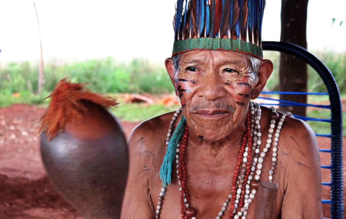 Papito Guarani trägt eine für die Guarani typische Feder-Kopfbedeckung und Ketten aus Samen, die in dem kleinen Rest Wald gesammelt wurden, der auf dem Land noch überleben konnte. Das Musikinstrument nennt sich mbaraka und wird für Rituale, Zeremonien und zum Tanzen verwendet.
