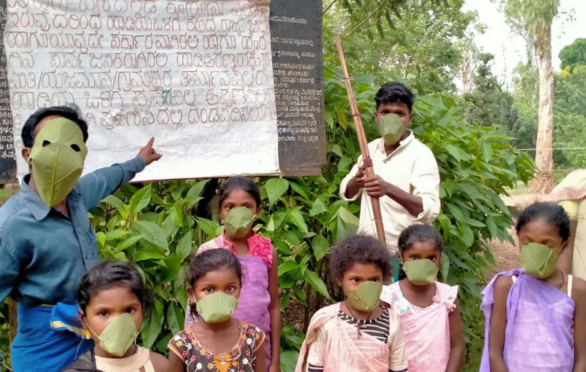 Jenu Kuruba wearing masks made of leaves from the sacred fig tree. They are standing in front of a notice telling outsiders to stay away from their hamlet, to protect them from Covid.